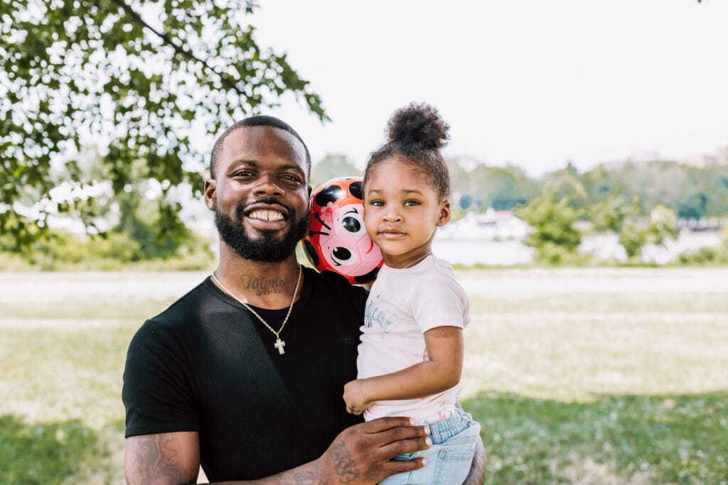 A smiling young man stands with his daughter (about 3 years old) in his arms. They are in a park with a field behind them.