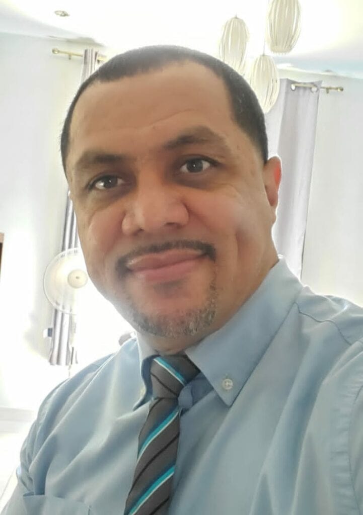 A man in a light blue dress shirt with a striped tie. He has short dark hair and a neat mustache and beard. He is smiling at the camera.
