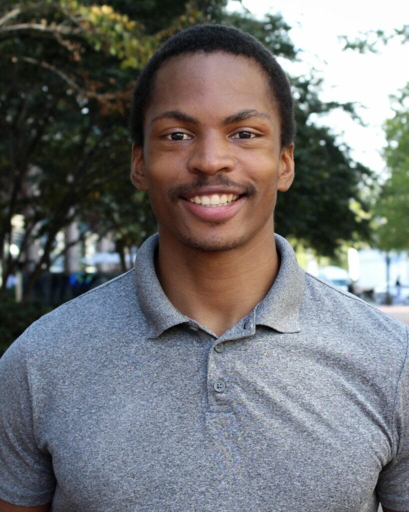 A young Black man smiling at the camera. He has short black hair and a mustache, and is wearing a gray polo shirt. There are trees visible in the background behind him.