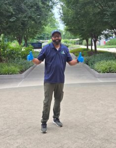 Man with a black beard stands in front of green bushes and trees. He's wearing a blue polo, khaki pants, and a blue hat. He has both hands out with giving a thumbs up. He is smiling.