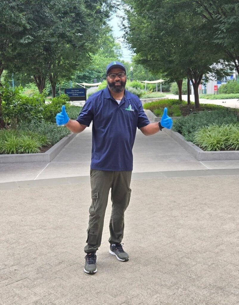 Man with a black beard stands in front of green bushes and trees. He's wearing a blue polo, khaki pants, and a blue hat. He has both hands out with giving a thumbs up. He is smiling.