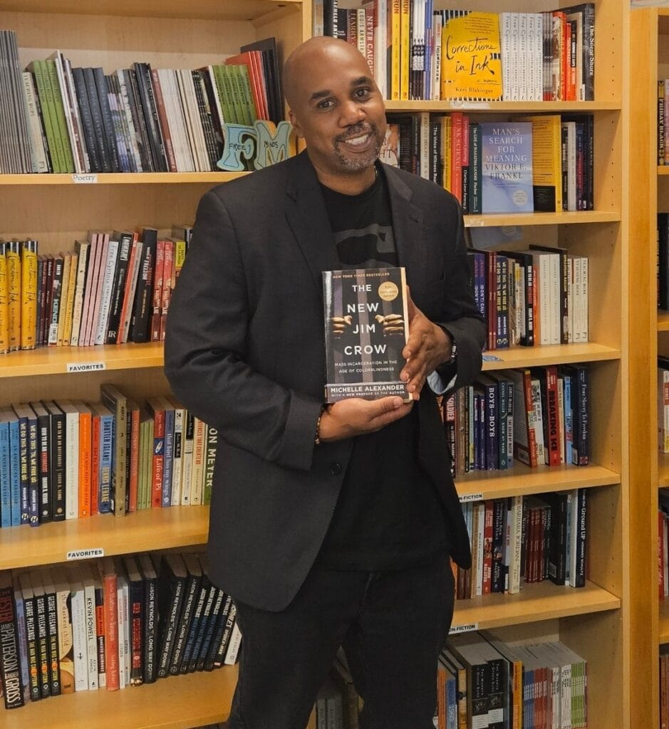 Man wearing a black shirt and blazer stands in front of a bookshelf. He is holding the book "The New Jim Crow" by Michelle Alexander