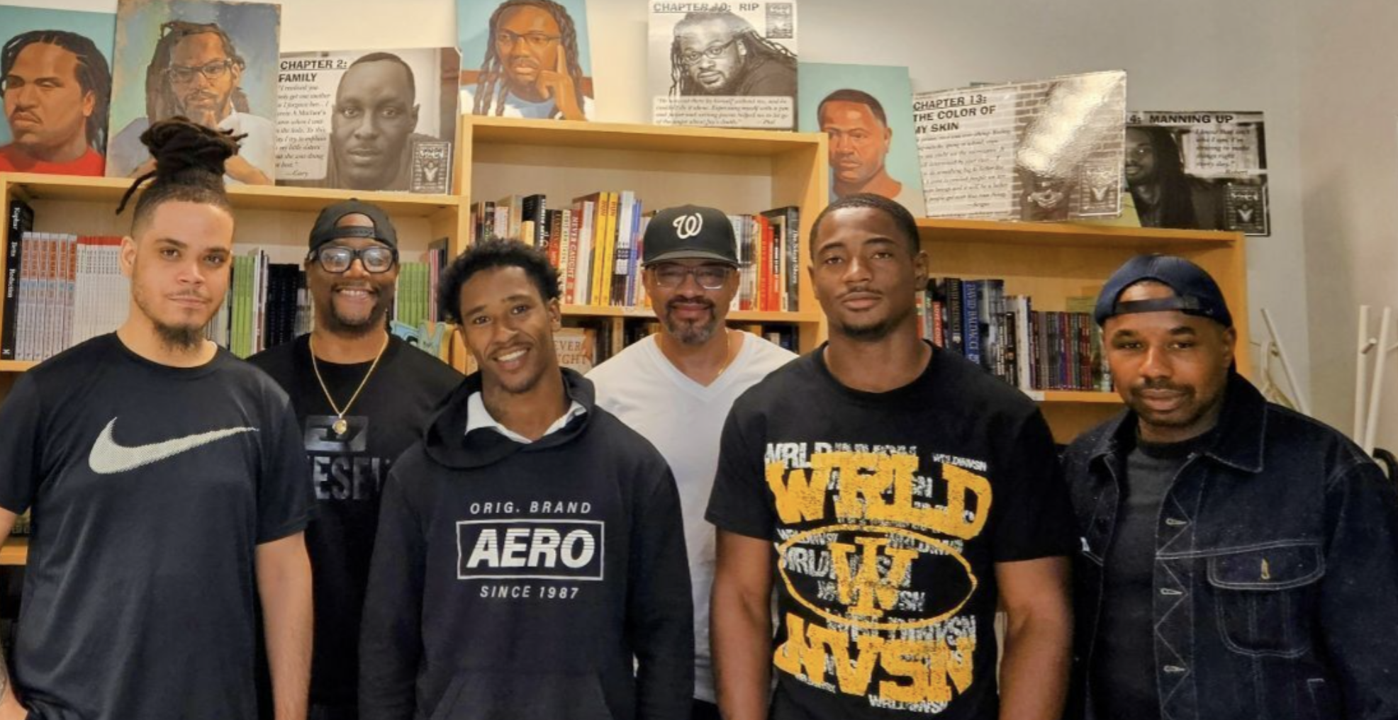 6 men standing in a row in front of a bookshelf